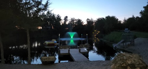 A serene pond at dusk with a green fountain, wooden docks, and trees reflecting in the water.