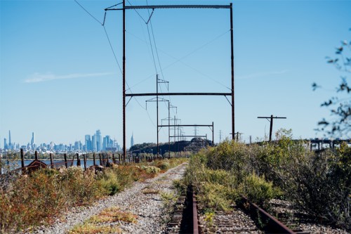 A view of railway tracks lined with power lines, leading towards a distant city skyline under a clear blue sky.