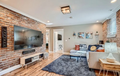 Cozy living room with exposed brick walls, a large TV, a sectional sofa, and a plush rug. Bright and inviting atmosphere.