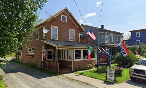 A cozy restaurant with a brown exterior, flags, and a welcoming porch, surrounded by greenery and a quiet street.