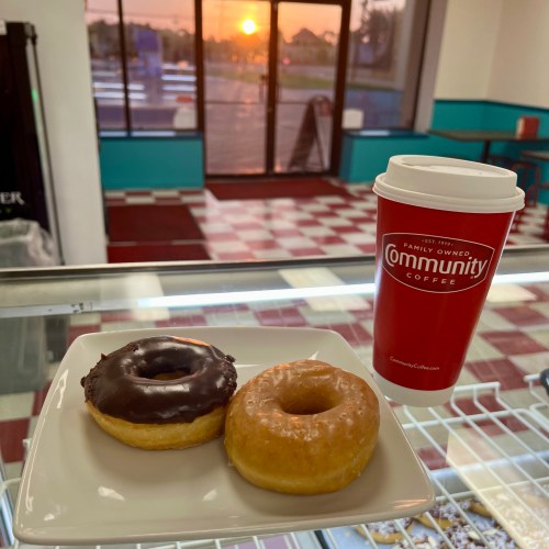 A plate with a chocolate donut and a glazed donut, next to a red coffee cup, with a sunset visible in the background.