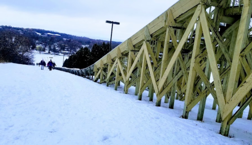 Two people walk up a snowy hill beside a wooden ski jump structure under a cloudy sky.