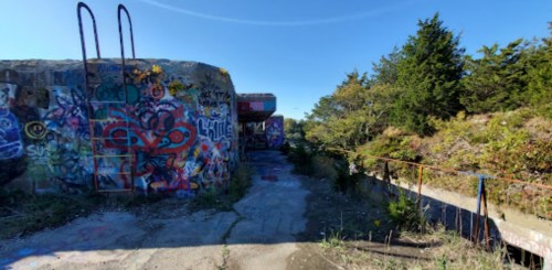 Abandoned structure with colorful graffiti, surrounded by greenery and a clear blue sky.