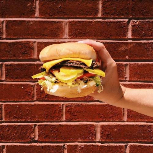 A hand holding a juicy burger with cheese, lettuce, tomato, and pickles against a brick wall background.