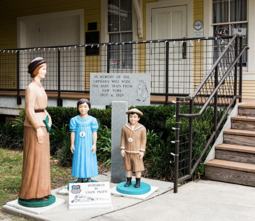 Three life-sized wooden figures of children in historical clothing stand outside a building, with a memorial stone nearby.
