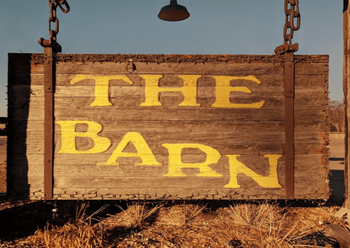 A weathered wooden sign hanging from chains, reading "THE BARN" in large yellow letters.