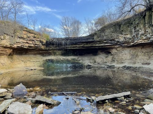 A serene landscape featuring a rocky cliff above a calm pond, surrounded by bare trees and a clear blue sky.