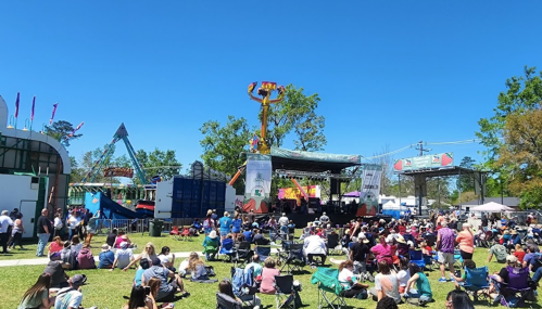 A lively outdoor festival scene with a crowd gathered in front of a stage, surrounded by rides and clear blue skies.