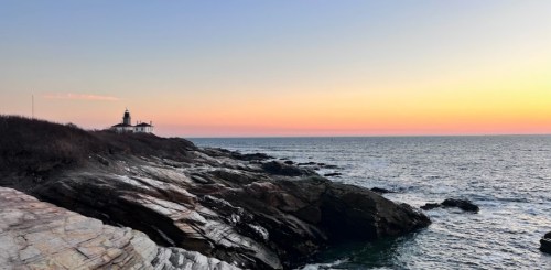 A serene coastal scene at sunset, featuring a lighthouse on rocky shores and calm ocean waters.