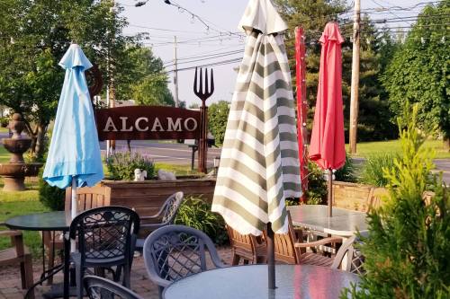 Outdoor seating area with colorful umbrellas and a sign reading "ALCAMO" in a landscaped setting.
