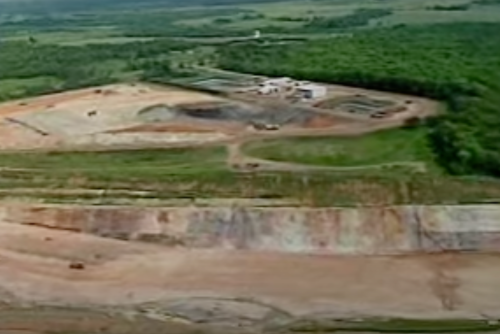 Aerial view of a large construction site with earthworks and machinery, surrounded by greenery and open land.