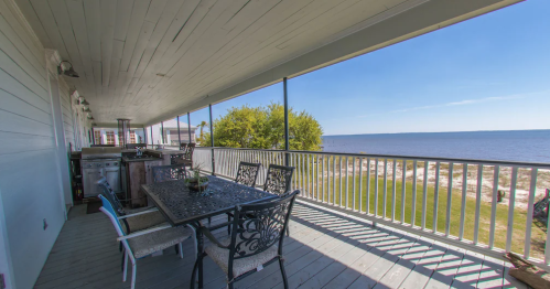 A spacious porch with a dining table overlooks a calm waterfront and green lawn under a clear blue sky.