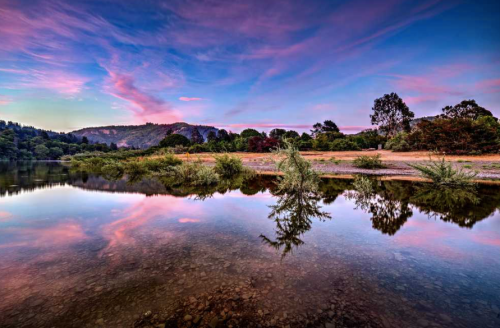 A serene river scene at sunset, with colorful clouds reflecting on the water and lush greenery along the shore.