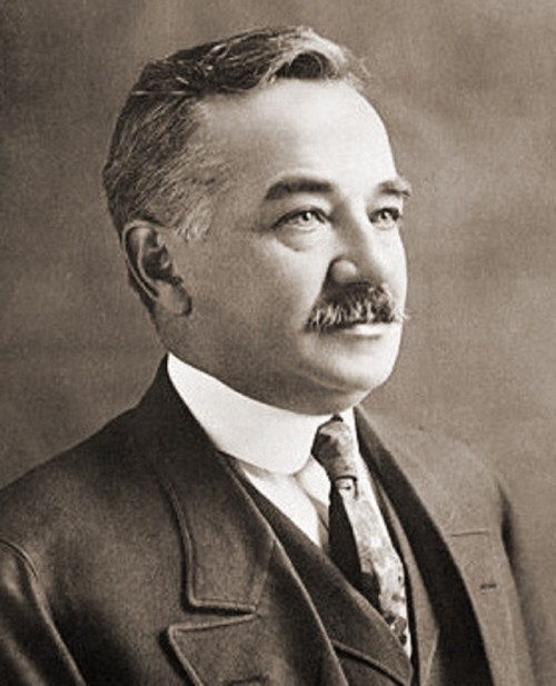 Black and white portrait of a man with a mustache, wearing a suit and tie, looking thoughtfully to the side.