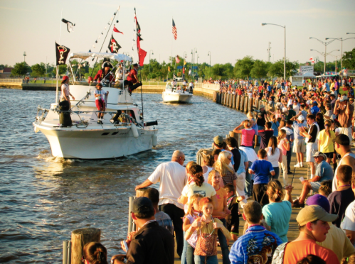 A crowded waterfront scene with boats, flags, and people enjoying a festive atmosphere by the water.
