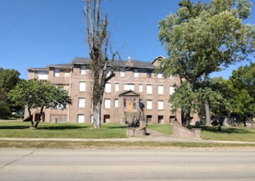 A large brick building with multiple windows, surrounded by trees and grass, set against a clear blue sky.