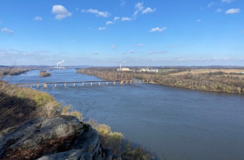 A scenic view of a river winding through a landscape with a bridge and distant industrial buildings under a blue sky.