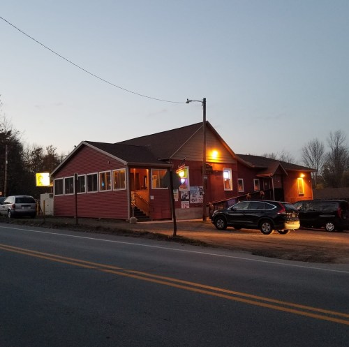 A red wooden building with a sloped roof, lit up at dusk, located by a road with parked cars nearby.