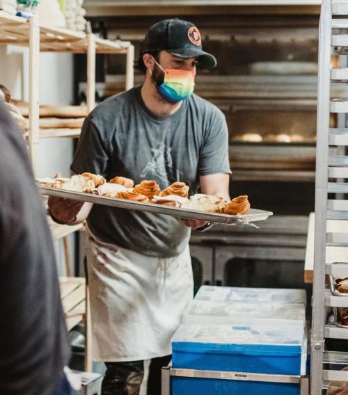 A baker wearing a rainbow mask holds a tray of pastries in a bakery, surrounded by shelves of baked goods.
