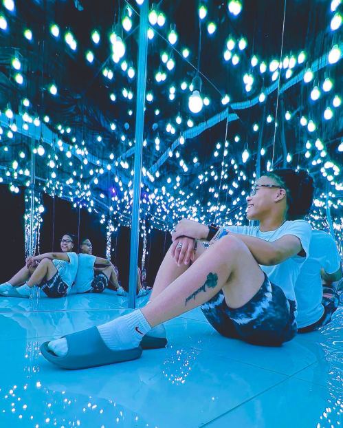 A person sits on the floor in a mirrored room filled with bright, glowing lights, smiling and enjoying the ambiance.
