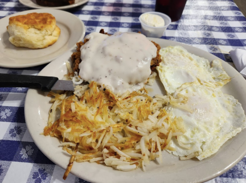 A plate of fried chicken smothered in gravy, hash browns, and two fried eggs, with a biscuit and drink in the background.