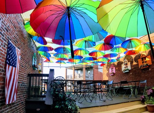 Colorful umbrellas hang overhead in a vibrant outdoor space, with a wooden deck, chairs, and an American flag on the wall.