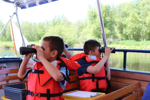 Two children in life jackets using binoculars on a boat, surrounded by greenery and calm water.