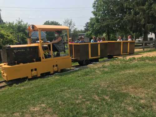 A yellow train with a driver pulls a group of children in a cart along a grassy track on a sunny day.