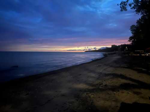 A serene beach at dusk, with calm waters and a colorful sky transitioning from blue to purple and orange.