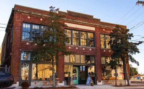 A brick building with large windows, surrounded by trees, featuring outdoor seating and a clear blue sky.