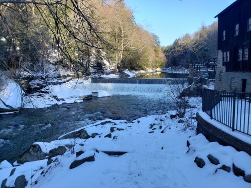 A serene winter scene featuring a river with a small waterfall, surrounded by snow-covered banks and trees.