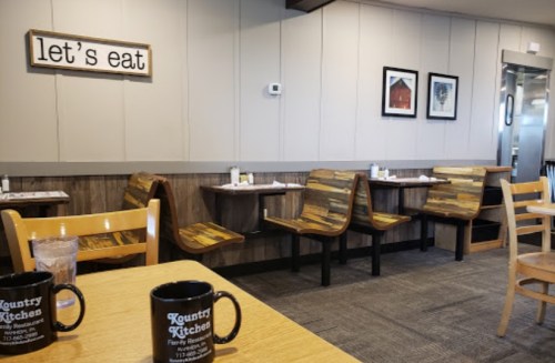 Cozy diner interior with wooden tables, chairs, and a "let's eat" sign on the wall. Coffee mugs on tables.