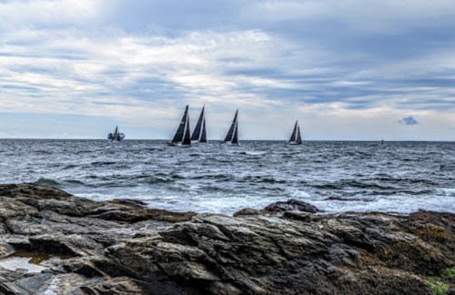 Sailboats with black sails navigate a choppy sea, with rocky shorelines and a cloudy sky in the background.