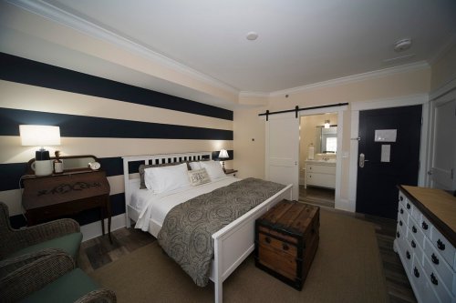 Cozy hotel room with a bed, striped walls, a wooden trunk, and a bathroom visible in the background.