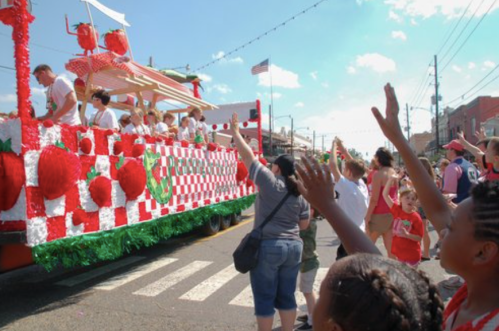 A festive parade float decorated with strawberries, with people waving and enjoying the celebration on a sunny day.