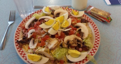A colorful salad with lettuce, tomatoes, mushrooms, cheese, and sliced hard-boiled eggs on a red-checkered plate.