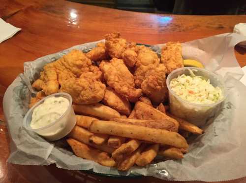 A platter of fried chicken tenders, French fries, coleslaw, and dipping sauce on a wooden table.