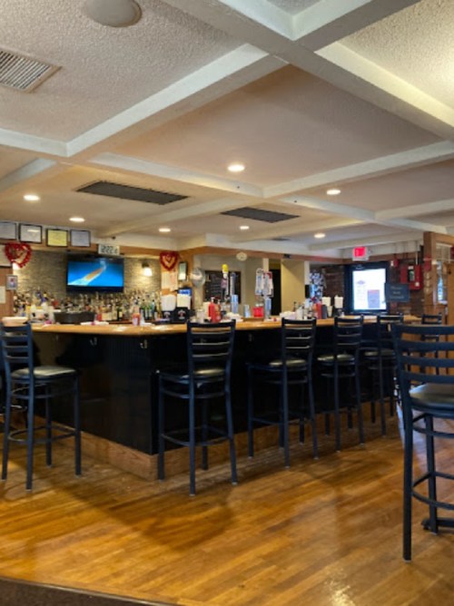 Interior of a bar with a wooden floor, black stools, and a counter displaying drinks and condiments. TV on the wall.