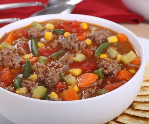 A hearty bowl of vegetable and beef soup with corn, carrots, and green beans, served with crackers on the side.