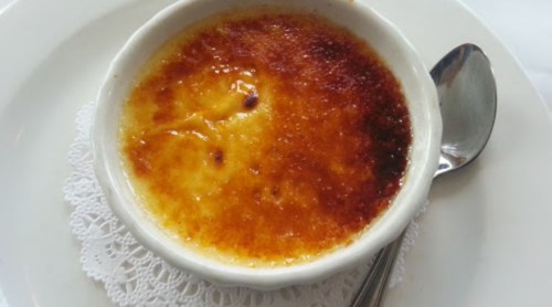 A creamy dessert in a white ramekin with a caramelized sugar top, served on a lace doily with a spoon beside it.