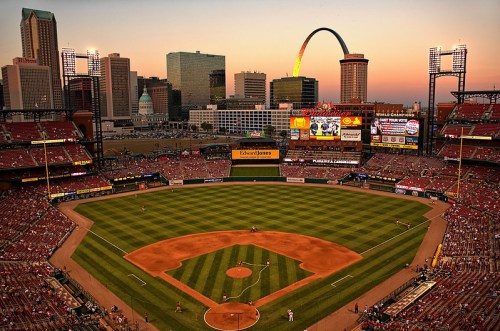 A baseball stadium at sunset, with a view of the city skyline and a colorful arch in the background.