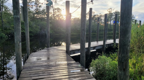 A wooden dock extends over calm water, surrounded by greenery, with a sunset in the background.