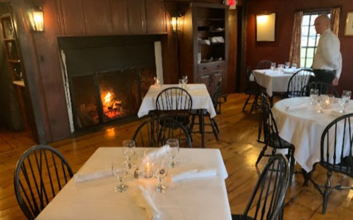 Cozy restaurant interior with wooden floors, tables set with white linens, and a fireplace glowing in the background.