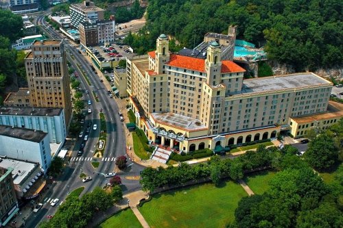 Aerial view of a historic hotel with red roofs, surrounded by greenery and a busy road in a scenic area.