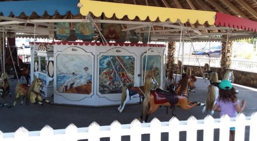 A vintage carousel with painted horses and colorful decorations, set in a shaded area with a child observing.