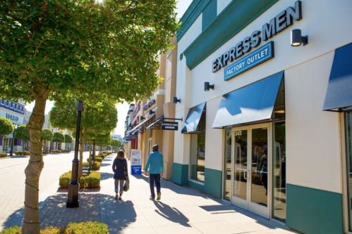 Two people walk past the Express Men Factory Outlet on a sunny day, with trees lining the sidewalk.