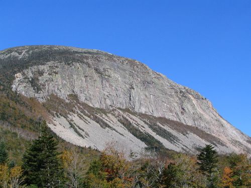 A large, rocky mountain rises against a clear blue sky, surrounded by trees and autumn foliage.