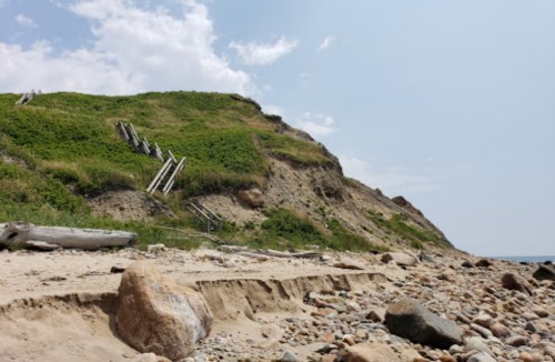 A sandy beach with a rocky shoreline and a grassy cliff, featuring wooden steps leading up the hill.