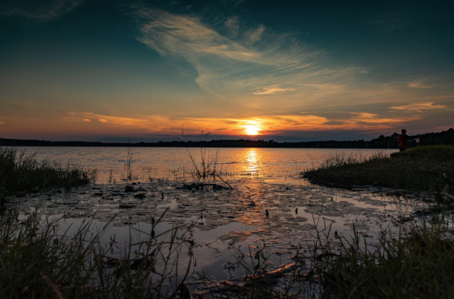 A serene sunset over a calm lake, with reflections on the water and grassy shores in the foreground.