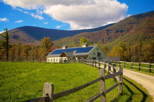 A scenic farm with a white house and solar panels, surrounded by green fields and mountains under a blue sky.